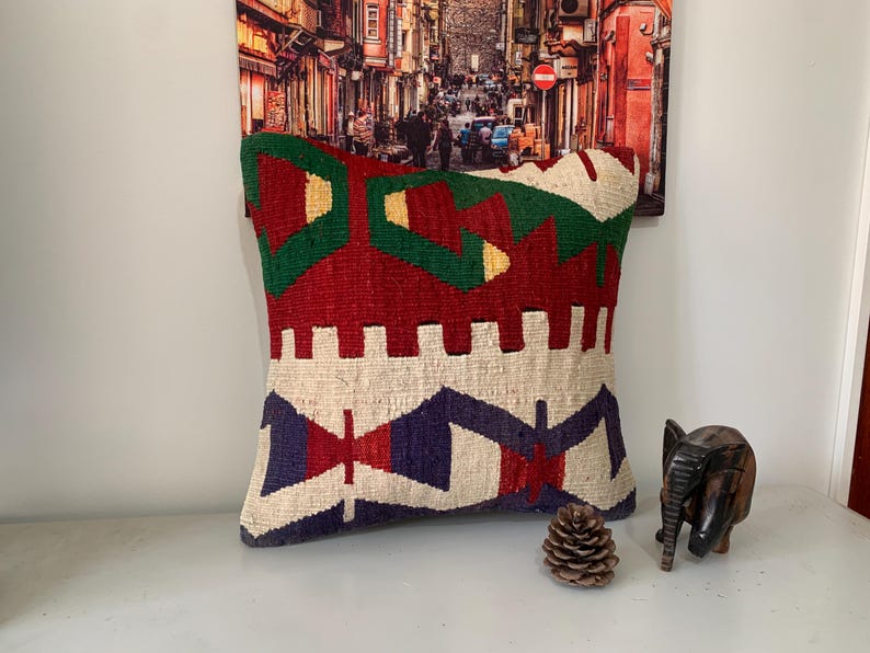 May include: A colorful throw pillow with a geometric pattern in shades of red, green, white, and blue. The pillow is made of woven fabric and has a fringed edge.