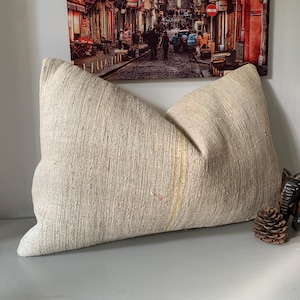 May include: A large, textured beige pillow with a soft, natural appearance. The pillow has a rectangular shape with a slight curve and a subtle vertical stripe. A small wooden elephant and a pine cone are next to the pillow.