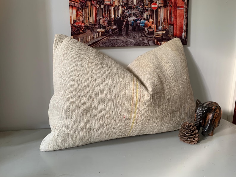 May include: A large, textured beige pillow with a soft, natural appearance. The pillow has a rectangular shape with a slight curve and a subtle vertical stripe. A small wooden elephant and a pine cone are next to the pillow.