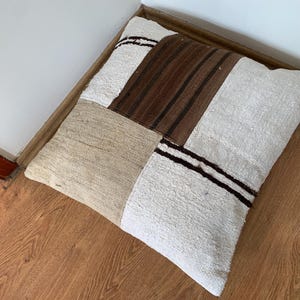 Puede incluir: Cojín cuadrado de patchwork en tonos neutros. Presenta secciones de tela crema, beige y marrón. Rayas marrones oscuras acentúan las secciones crema. El cojín está colocado sobre un suelo de madera.