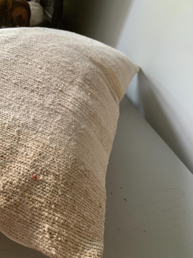 May include: A beige woven pillow with a textured surface. The pillow has a rectangular shape and appears to be made of a natural fiber. The pillow is placed against a white wall.