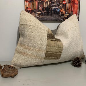 May include: A white and brown patchwork pillow with a textured woven fabric. The pillow is on a light brown surface with a small wooden elephant figurine and a pine cone.