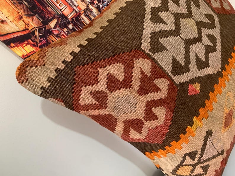 May include: A brown, orange and beige patterned throw pillow with a geometric design. The pillow is made of woven fabric and has a fringed edge.