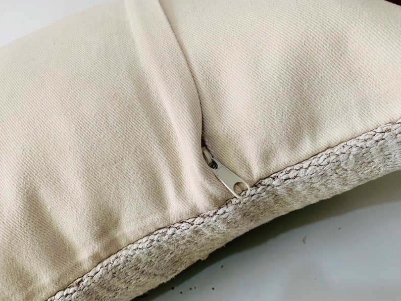 May include: A close-up of a cream-coloured pillow with a zipper closure. The pillow has a textured fabric border.
