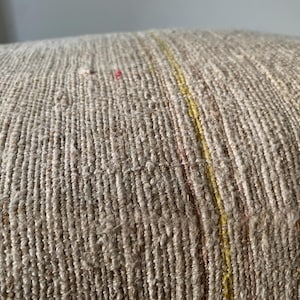 May include: Close-up of a textured, beige fabric with vertical lines. A thin yellow stripe runs vertically through the center, with hints of pink. The fabric appears to be woven, possibly for a pillow or upholstery.