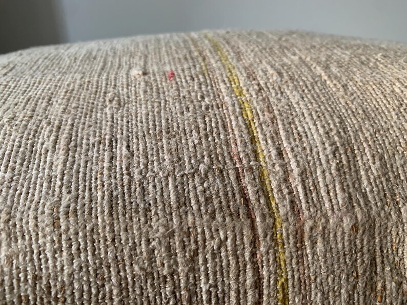 May include: Close-up of a textured, beige fabric with vertical lines. A thin yellow stripe runs vertically through the center, with hints of pink. The fabric appears to be woven, possibly for a pillow or upholstery.