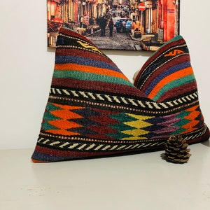 May include: A colorful, patterned pillow with a geometric design in shades of red, orange, yellow, green, blue, and purple. The pillow is made of woven fabric and has a fringed edge.