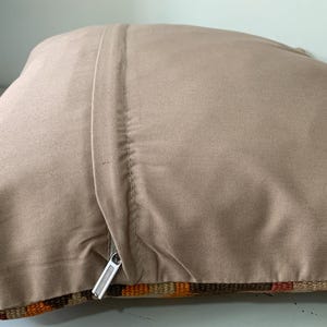 May include: A beige pillow cover with a zipper closure. The cover is made of a soft, textured fabric and has a subtle sheen. The pillow cover is shown folded in half, revealing the zipper and the back side of the fabric.