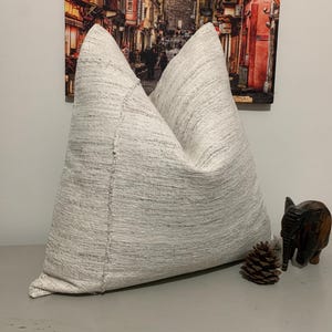 May include: A large, textured, off-white decorative pillow. The pillow is made of a woven fabric with a subtle pattern. A small wooden elephant figurine and a pine cone are next to the pillow.