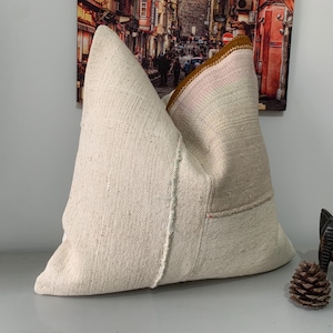 May include: A white and cream colored pillow with a brown and pink trim. The pillow is made of a woven fabric and has a textured surface.