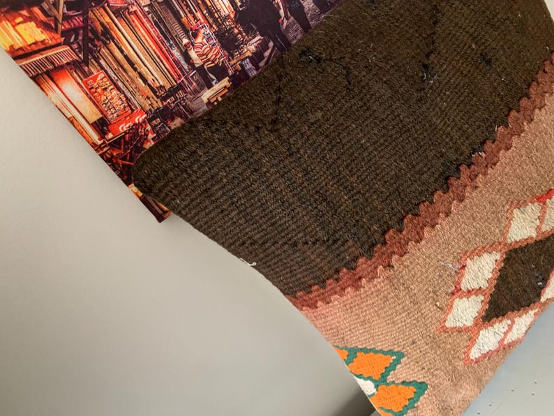 May include: A brown, beige, and orange patterned throw pillow with a geometric design. The pillow is made of woven fabric and has a fringed edge.