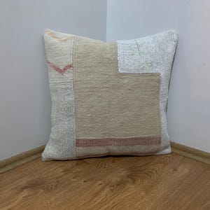 May include: Square decorative pillow with a patchwork design. The pillow features a mix of neutral tones, including beige, cream, and light pink, with a textured appearance. The pillow is placed in a corner against a white wall and wooden floor.