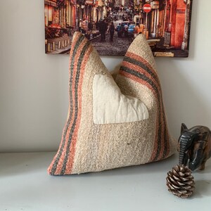 May include: A beige and orange striped pillow with a white patch. The pillow is placed on a white surface, with a wooden elephant figurine and a pine cone nearby. A framed picture hangs in the background.