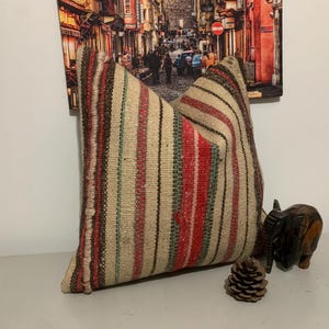 Könnte beinhalten: Ein beiges Kissen mit vertikalen Streifen in Rot, Grün und Braun. Das Kissen besteht aus einem gewebten Stoff. Ein kleiner Elefant aus Holz und ein Tannenzapfen liegen neben dem Kissen.