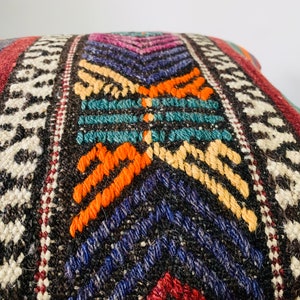 May include: A colorful woven pillow with a geometric pattern in shades of red, orange, yellow, blue, green, and white. The pillow is made of a thick, textured fabric and has a fringe border.