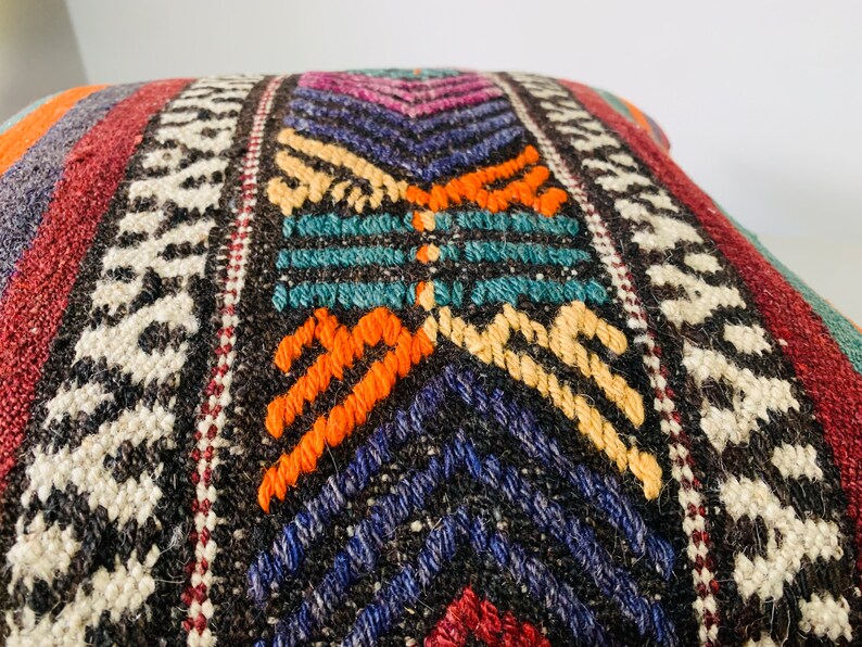 May include: A colorful woven pillow with a geometric pattern in shades of red, orange, yellow, blue, green, and white. The pillow is made of a thick, textured fabric and has a fringe border.