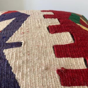 May include: A close-up of a decorative pillow with a geometric pattern in shades of red, white, blue, and green. The pillow is made of a woven fabric with a textured surface.