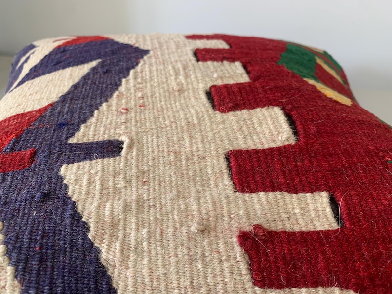 May include: A close-up of a decorative pillow with a geometric pattern in shades of red, white, blue, and green. The pillow is made of a woven fabric with a textured surface.