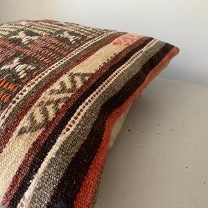 May include: Close-up of a decorative pillow with a woven design. The pillow features a mix of colors, including brown, orange, beige, and white. The pattern includes geometric shapes and stripes, creating a textured appearance. The pillow is likely used for home decor.