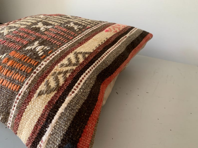 May include: Close-up of a decorative pillow with a woven design. The pillow features a mix of colors, including brown, orange, beige, and white. The pattern includes geometric shapes and stripes, creating a textured appearance. The pillow is likely used for home decor.