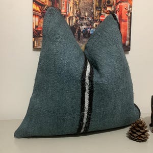 Puede incluir: Una almohada de tiro azul turquesa con rayas blancas y negras en el centro. La almohada está hecha de una tela suave y texturizada.