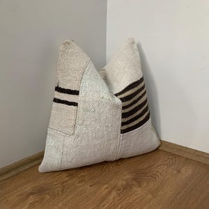 May include: A beige cushion with a patchwork design. The cushion features dark brown stripes and rectangular patches. The cushion is made of a textured fabric and is placed in the corner of a room with wooden flooring.