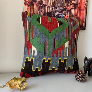 May include: A colorful throw pillow with a geometric pattern in shades of red, green, blue, and black. The pillow is made of woven fabric and has a fringed edge.