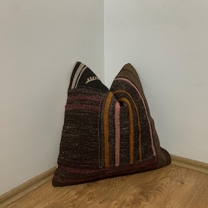 May include: A decorative pillow with a patchwork design. The pillow features a mix of brown, black, and red fabrics. Vertical stripes in pink and orange adorn a section of the pillow. The pillow is placed in a corner against a white wall and wooden floor.