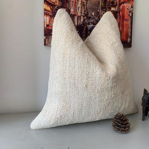 May include: A large, off-white, square cushion with a textured woven fabric. The cushion is sitting on a white surface with a small pine cone and a wooden elephant figurine.