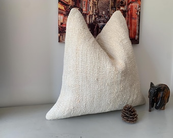 May include: A large, off-white, square pillow with a textured woven fabric. The pillow is sitting on a white surface with a small pine cone and a wooden elephant figurine.