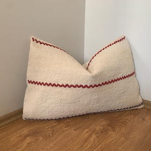 Puede incluir: Cojín rectangular color crema con costuras rojas en zigzag en los bordes y una línea horizontal. El cojín está hecho de una tela texturizada y se coloca en una esquina contra una pared blanca y un suelo de madera.
