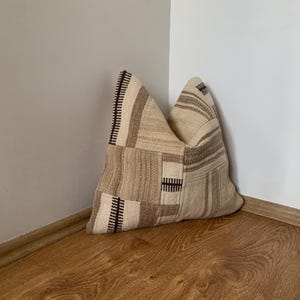 Może przedstawiać: Beżowo-brązowa poduszka patchwork z geometrycznym wzorem. Poduszka wykonana jest z tkanej tkaniny i ma teksturowany wygląd. Umieszczona jest w rogu pomieszczenia z drewnianą podłogą i białymi ścianami.