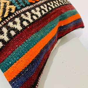 May include: A colorful striped pillow cover with a geometric pattern in the top corner. The pillow cover is made of woven fabric and has a variety of colors, including red, orange, blue, green, and white.