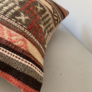 May include: Close-up of a decorative pillow with a woven design. The pillow features a mix of colors, including brown, red, beige, and pink. The pattern includes geometric shapes and stripes, creating a textured appearance.