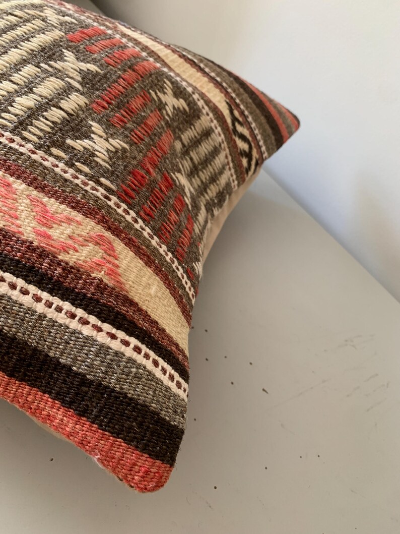 May include: Close-up of a decorative pillow with a woven design. The pillow features a mix of colors, including brown, red, beige, and pink. The pattern includes geometric shapes and stripes, creating a textured appearance.