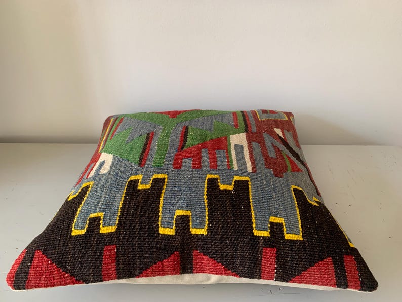 May include: A colorful, geometric patterned throw pillow with a blue, green, red, black, and yellow design. The pillow is made of woven fabric and has a white backing.