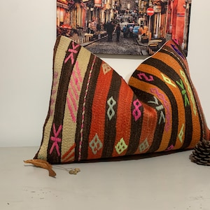 May include: A colorful pillow cover with a geometric pattern in shades of brown, orange, pink, and green. The pillow cover is made of woven fabric and has a fringe trim.