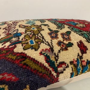 Puede incluir: Una almohada decorativa con un fondo de color crema y un estampado floral colorido. El estampado presenta tonos rojos, azules, verdes y marrones. La almohada tiene un borde blanco.