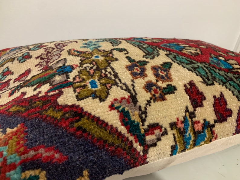 Puede incluir: Una almohada decorativa con un fondo de color crema y un estampado floral colorido. El estampado presenta tonos rojos, azules, verdes y marrones. La almohada tiene un borde blanco.
