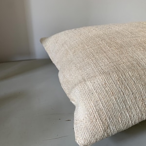 May include: A rectangular, off-white pillow with a textured, woven fabric. The pillow is angled, showing its corner and side. The fabric has a natural, rustic appearance, suggesting a handmade or vintage item. The pillow is likely for home decor.