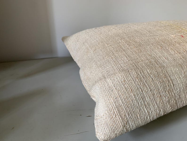 May include: A rectangular, off-white pillow with a textured, woven fabric. The pillow is angled, showing its corner and side. The fabric has a natural, rustic appearance, suggesting a handmade or vintage item. The pillow is likely for home decor.