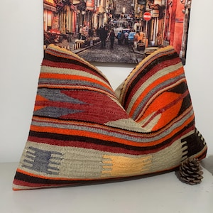 May include: A large, rectangular cushion with a colourful, striped pattern in shades of red, orange, beige, and grey. The cushion is made of woven fabric and has a fringed edge.