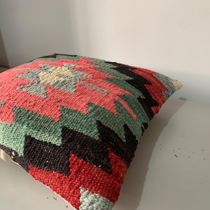 May include: A colorful throw pillow with a geometric pattern in shades of red, green, black, and white. The pillow is made of woven fabric and has a soft, textured surface.