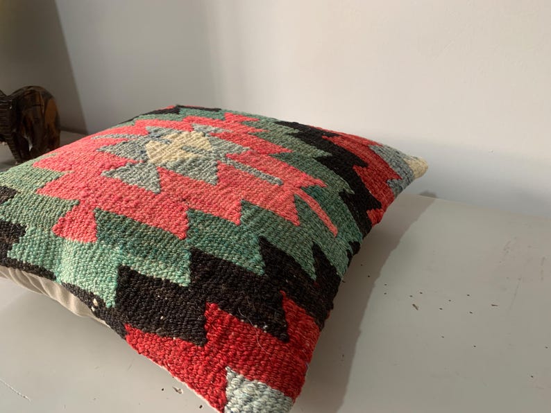 May include: A colorful throw pillow with a geometric pattern in shades of red, green, black, and white. The pillow is made of woven fabric and has a soft, textured surface.