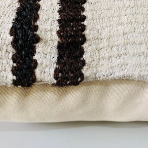 May include: Close-up of a cream-coloured throw pillow with black and brown stripes. The pillow has a textured woven fabric and a plain cream-coloured backing.