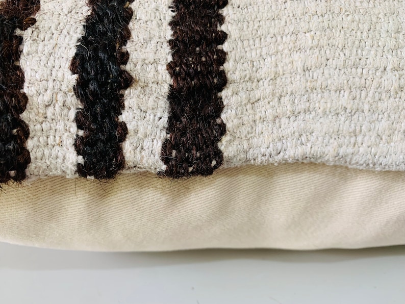 May include: Close-up of a cream-coloured throw pillow with black and brown stripes. The pillow has a textured woven fabric and a plain cream-coloured backing.