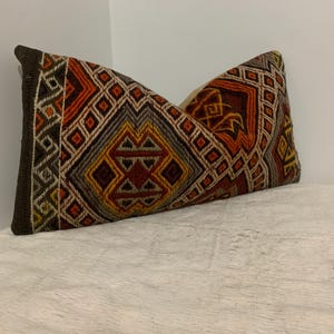 May include: Rectangular decorative pillow with a geometric pattern in shades of brown, orange, yellow, and gray. The pillow has a dark brown border and is placed on a white surface. The pillow's design features diamond and rectangular shapes.