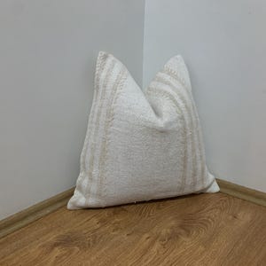 May include: A white pillow with vertical beige stripes, resting in a corner. The pillow has a textured appearance and is made of a woven fabric. The pillow is a decorative item.