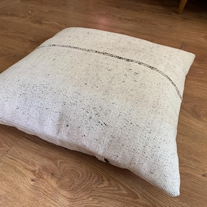 May include: Square, off-white floor pillow with a textured surface and black flecks. A dark, thin line runs across the center. The pillow is placed on a wooden floor.