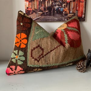 May include: A decorative pillow with a brown, beige, and colorful floral pattern. The pillow features orange, pink, and green flowers on a dark brown background. The pillow is placed on a white surface.
